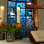 alter mit Kreuz, aufgeschlagener Bibel, brennenden Kerzen und Blumenschmuck vor blauem Kirchenfenster